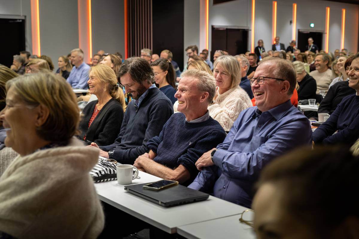 Et lattermildt publikum i en stor sal på InSchool Live. De sitter med mindre pulter og arbeidssaker foran seg.