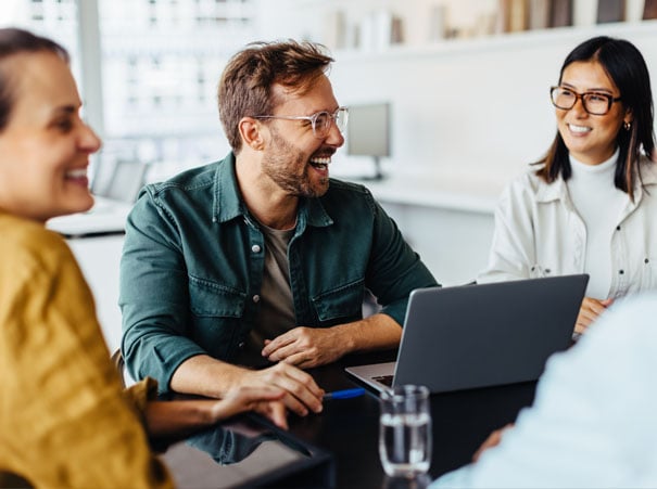 Engasjerte kollegaer i møte på et moderne kontor, der en mann ler foran en bærbar datamaskin. Bildet illustrerer hvordan InSchools kompetanseavtale legger til rette for fleksibel opplæring, rådgivning og effektivt samarbeid.