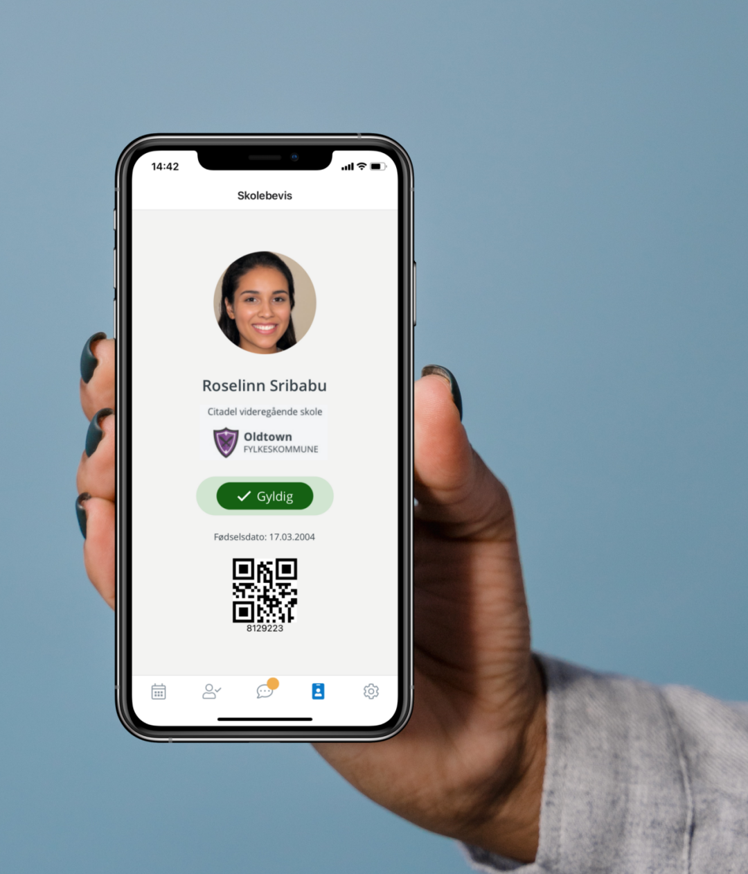 En smarttelefon holdes opp foran kamera, og viser et digitalt skolebevis. Skolebeviset har et elevbilde, informasjon om skole og fylkeskommune, gyldighetsbevis og en strekkode.